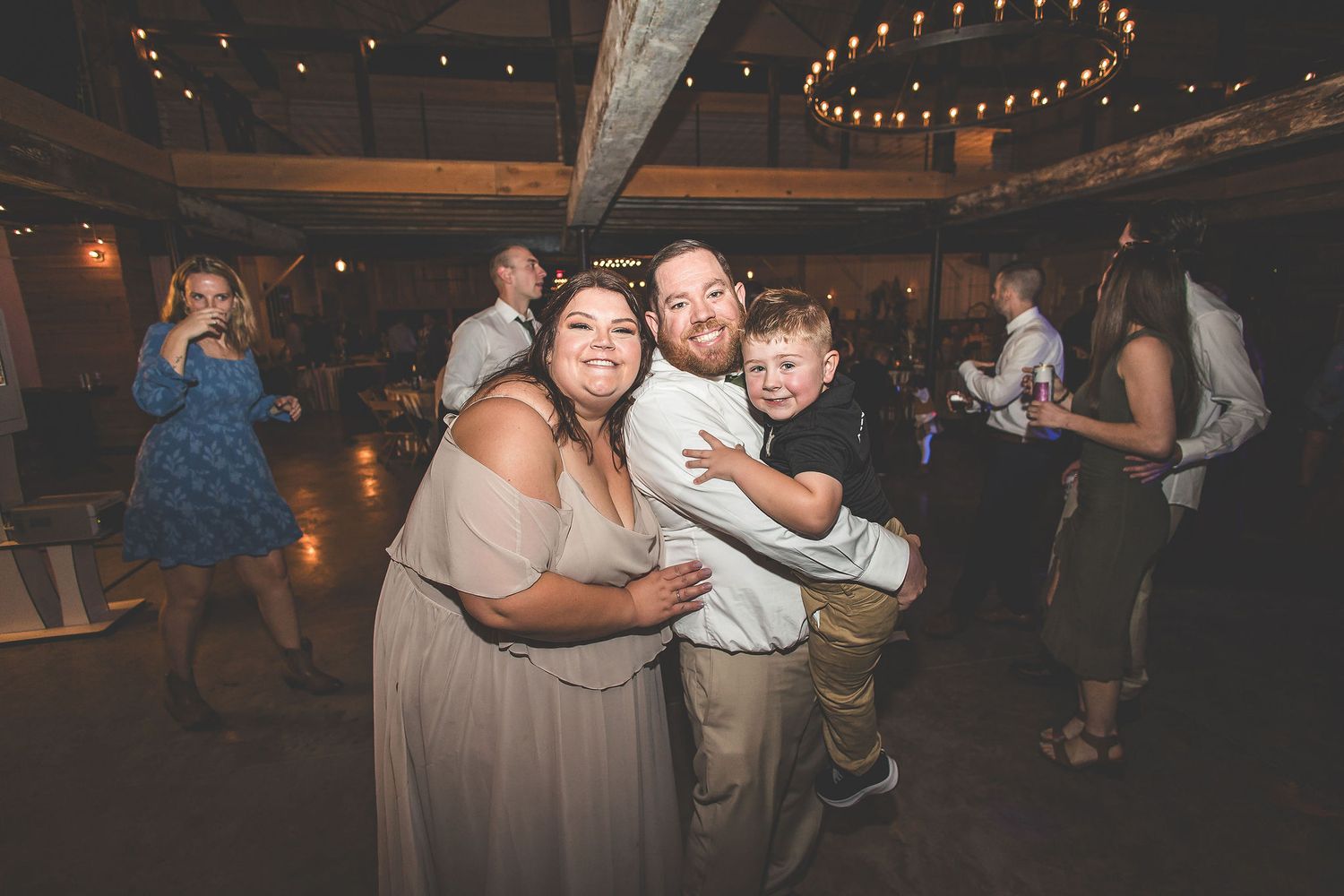 Guests dance and celebrate at a wedding reception under string lights in a rustic venue.