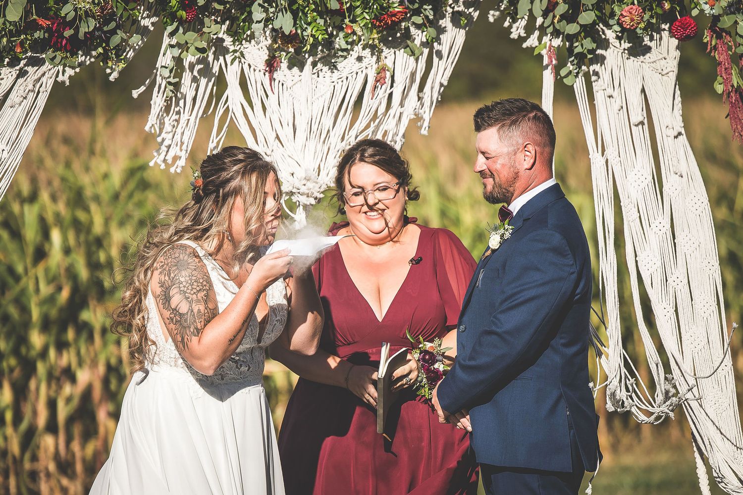 Wedding party in navy and burgundy attire gathered under floral arch with macrame details.