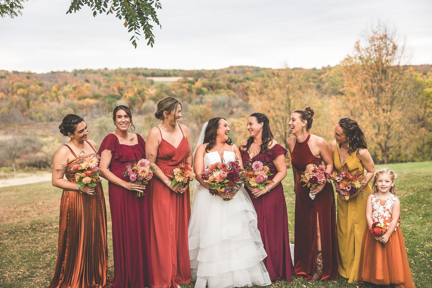 Bridal party wearing warm autumn colors stands together on a grassy hillside during fall.