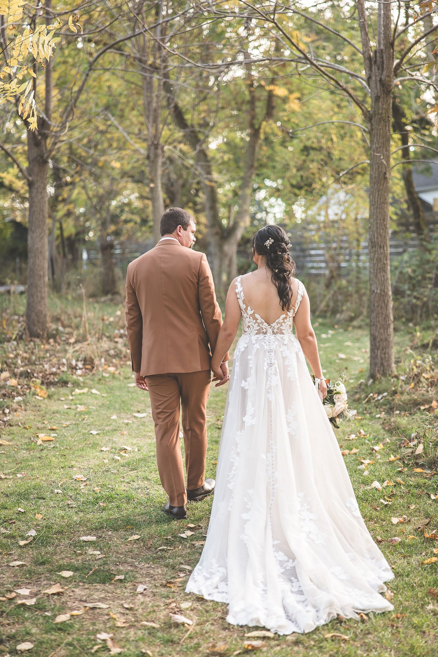 Couple in wedding attire walking away on leaf-covered path through autumn woods.