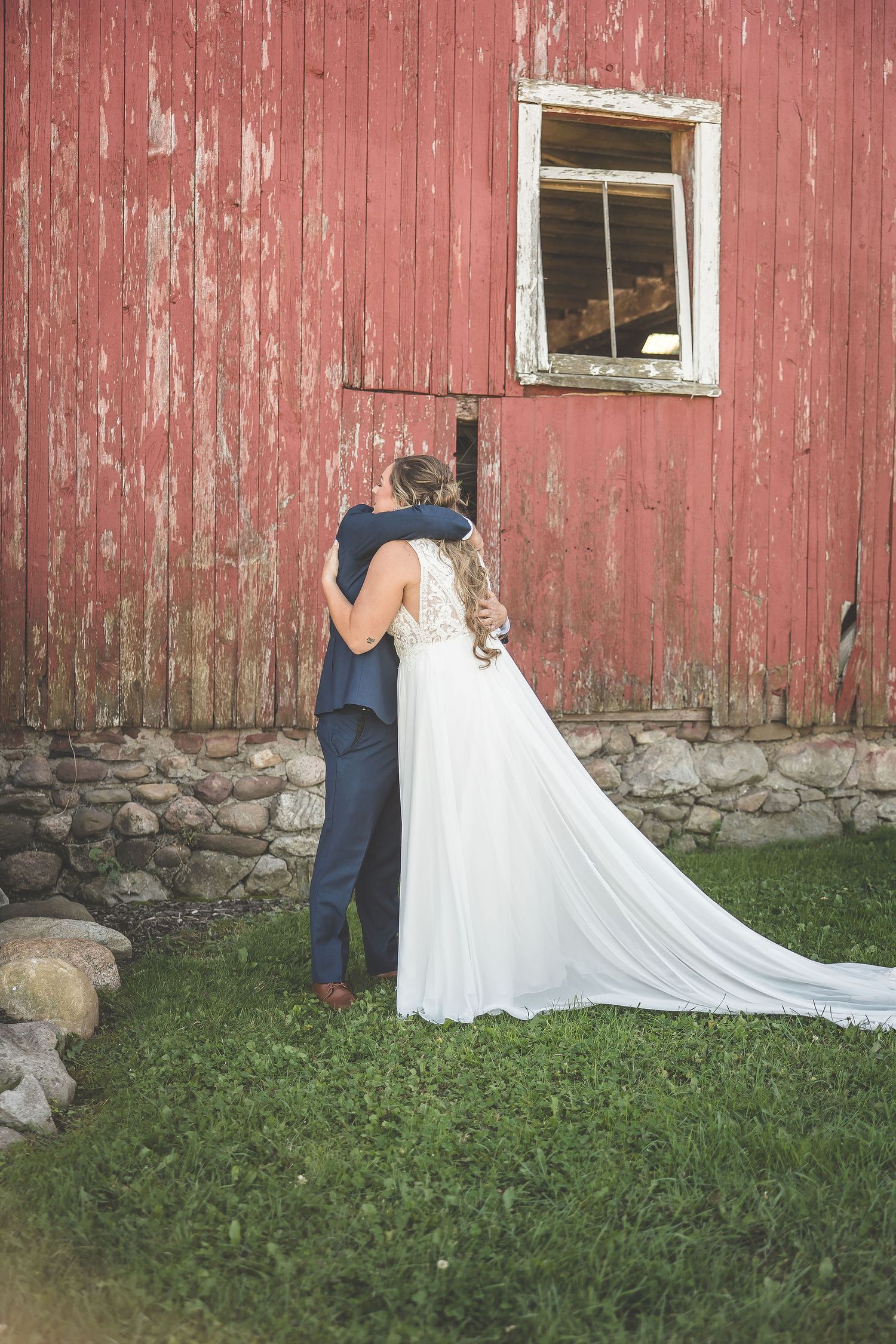 Bride's flowing white train trails beautifully against rustic red barn backdrop.