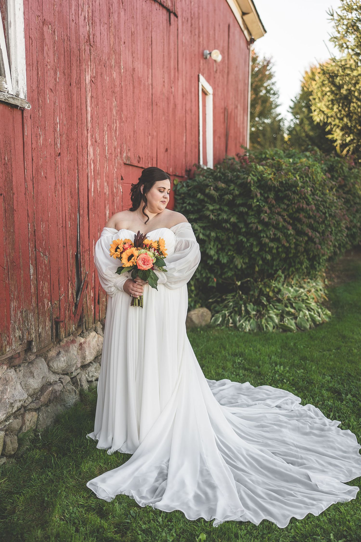 Bride in flowing white dress with train poses against red barn in autumn wedding setting.
