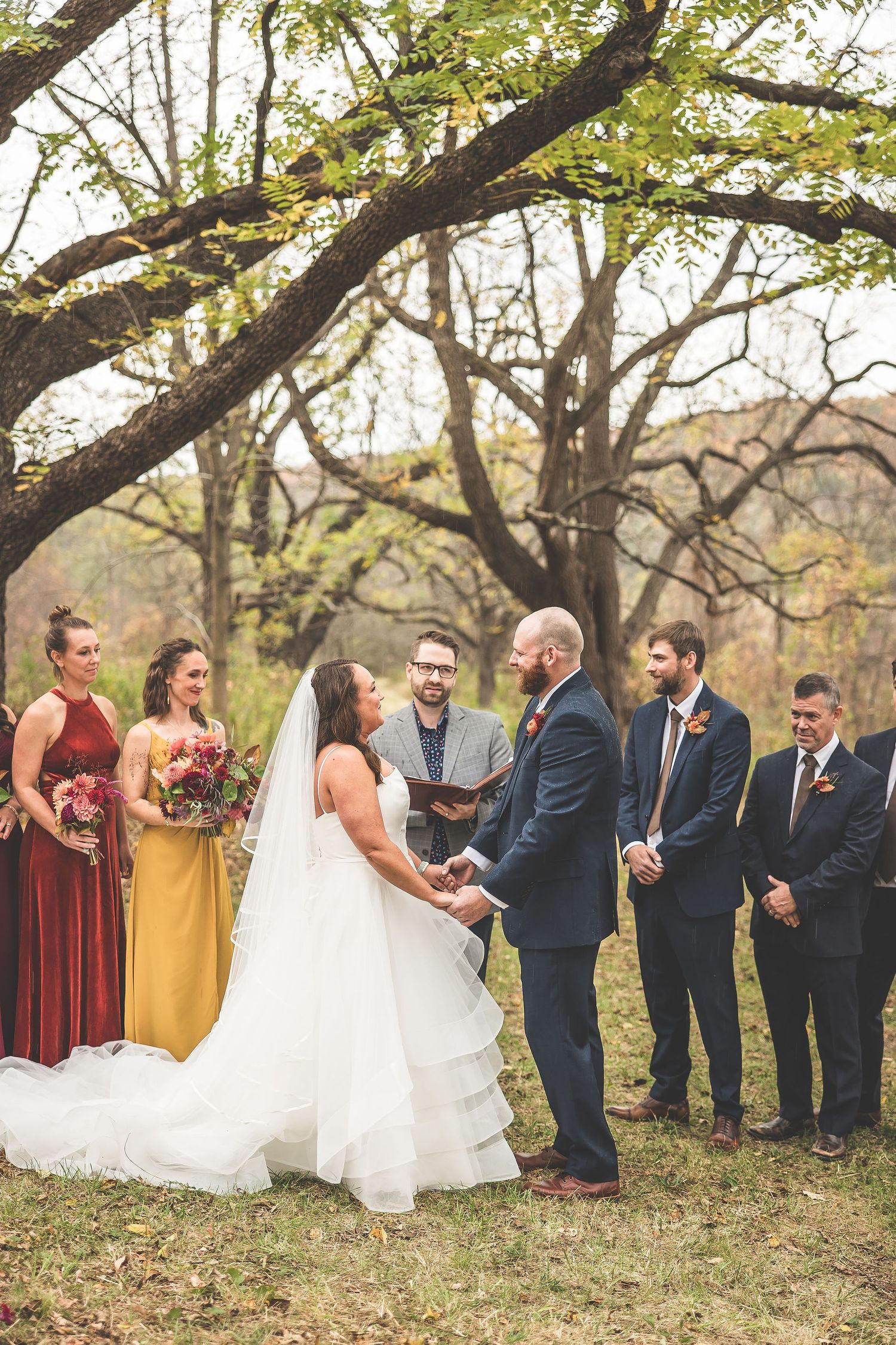 Autumn outdoor wedding ceremony with bridal party in burgundy and navy attire under trees.