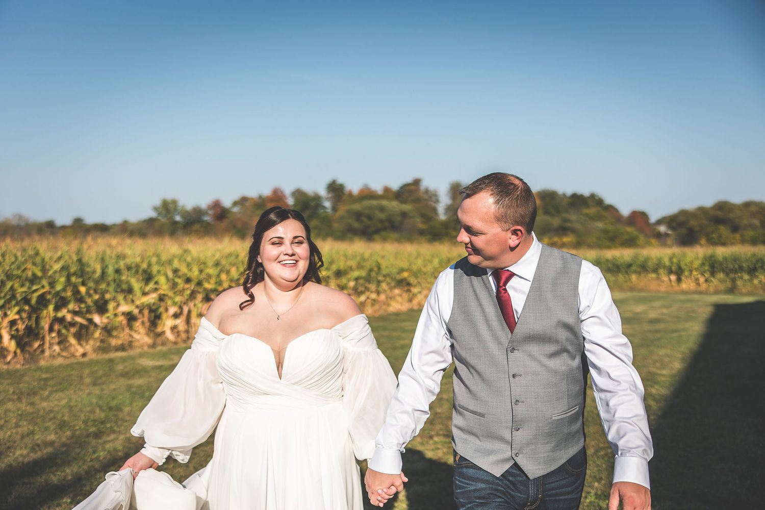 Newlyweds walking through a sunny field with dried corn stalks in the background.
