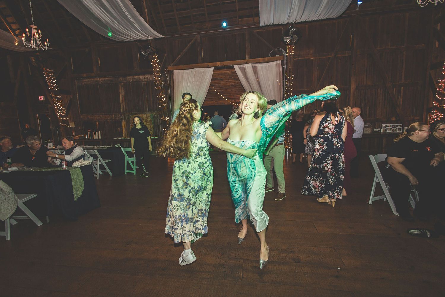 Two dancers perform an energetic move on a wooden dance floor under green lighting at a reception.