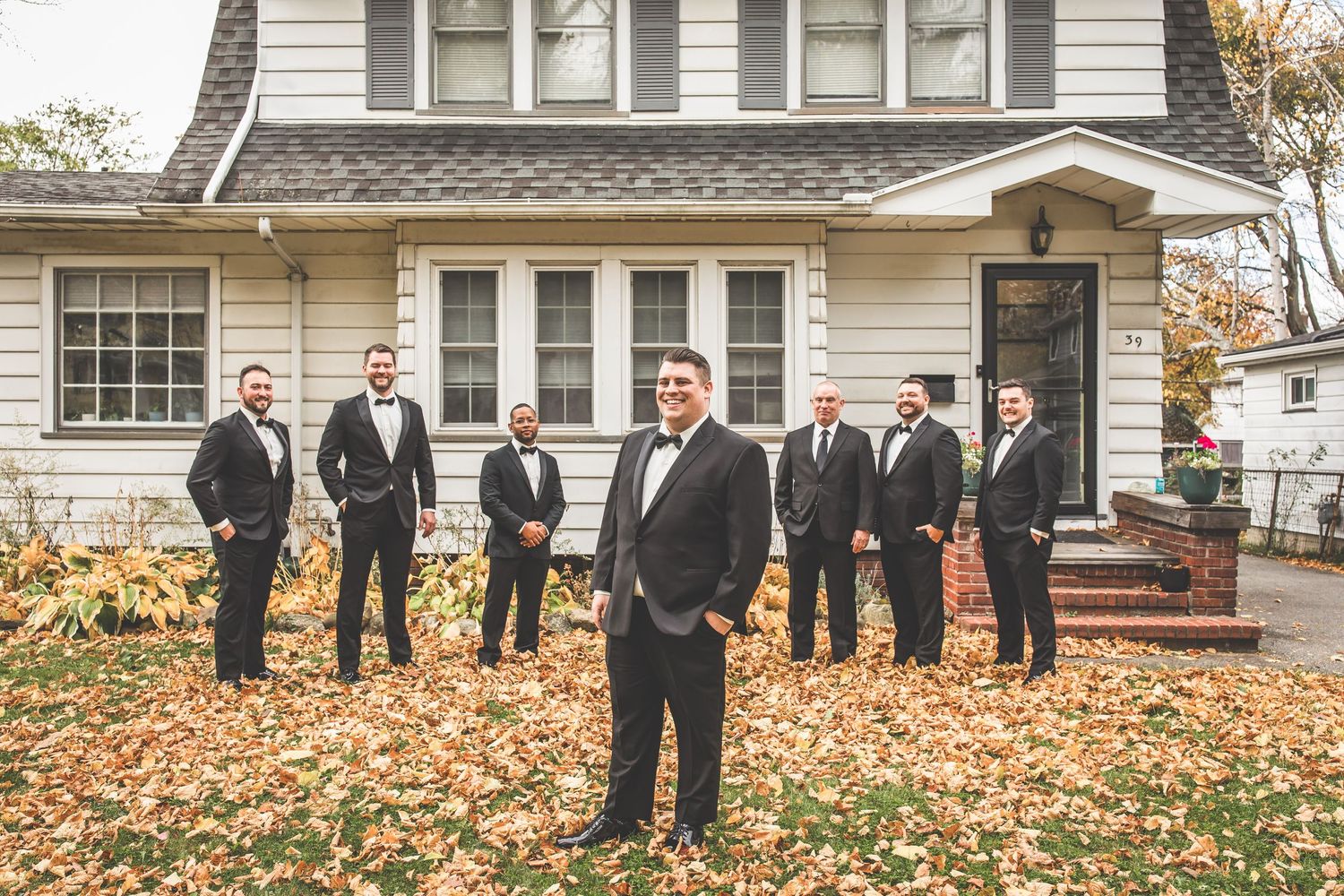 A formal wedding party poses outside a classic white house with fall foliage on the ground.