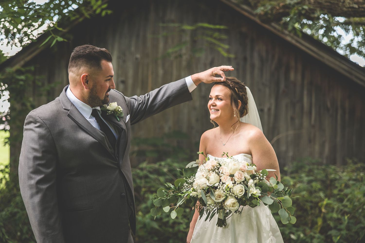 Newlyweds share an intimate moment in front of a weathered barn surrounded by lush greenery.