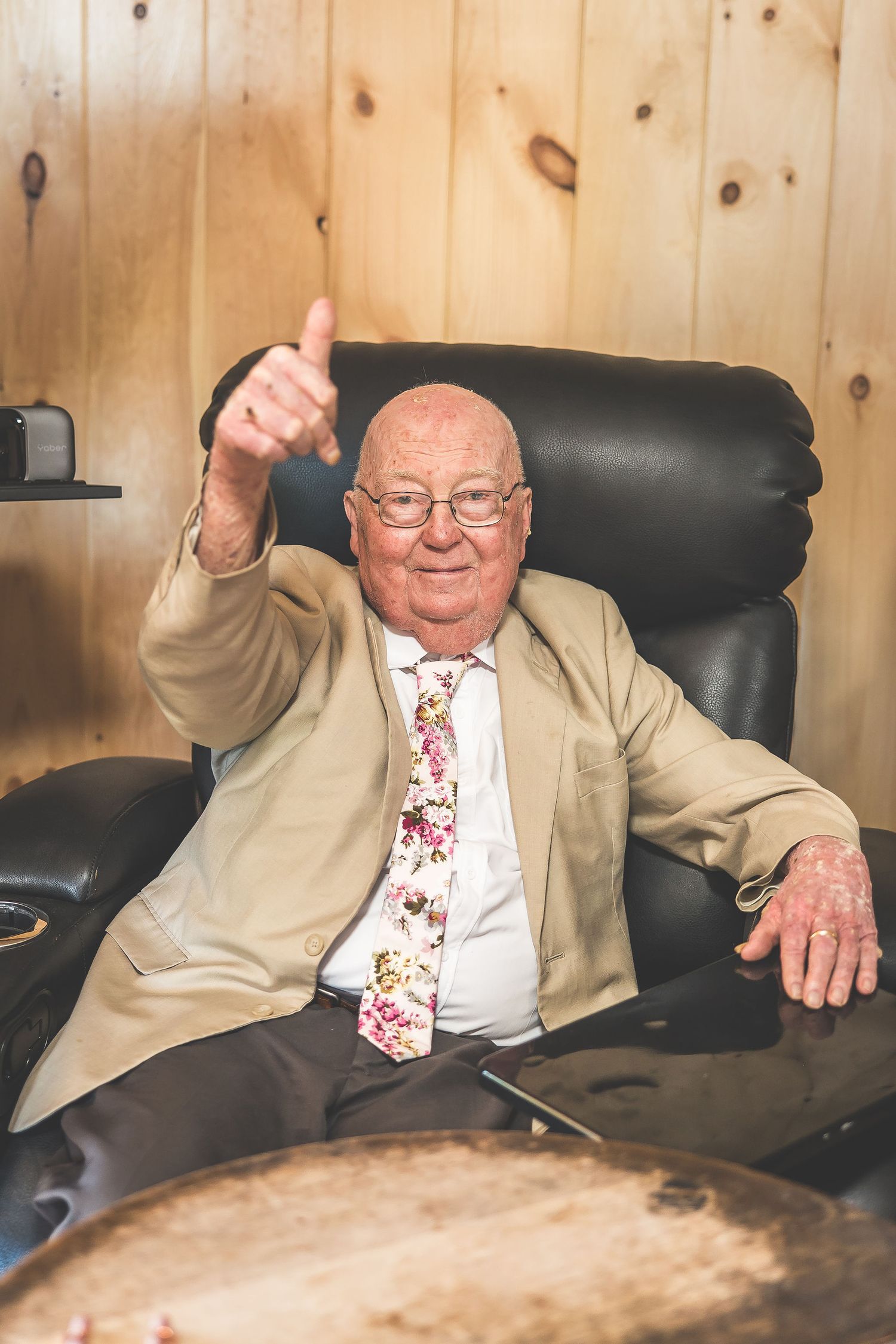 Elderly person in a beige suit and floral tie gestures animatedly while seated in a black leather chair.