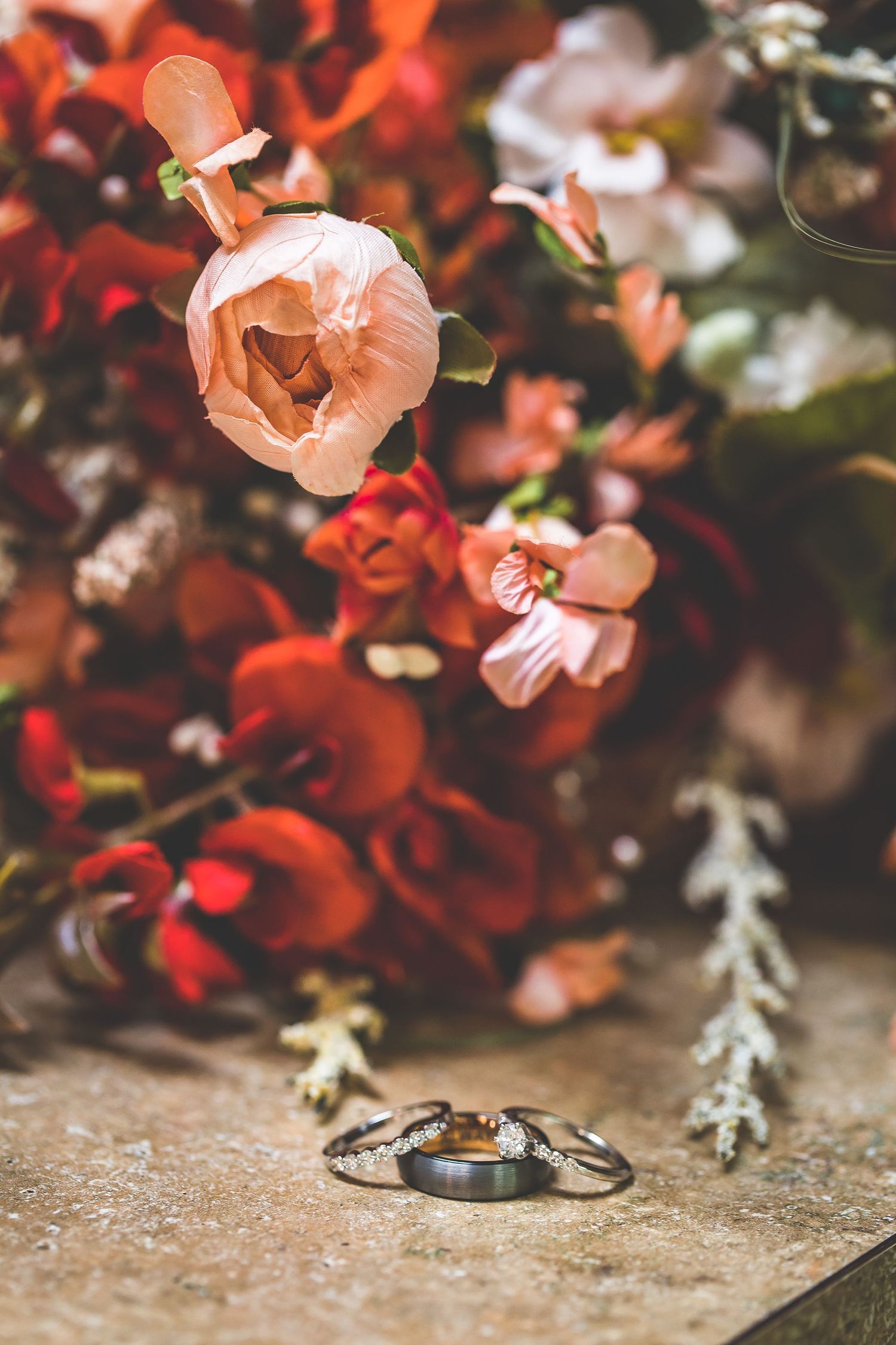 Wedding rings arranged beneath pink and red flowers with artistic bokeh effect.
