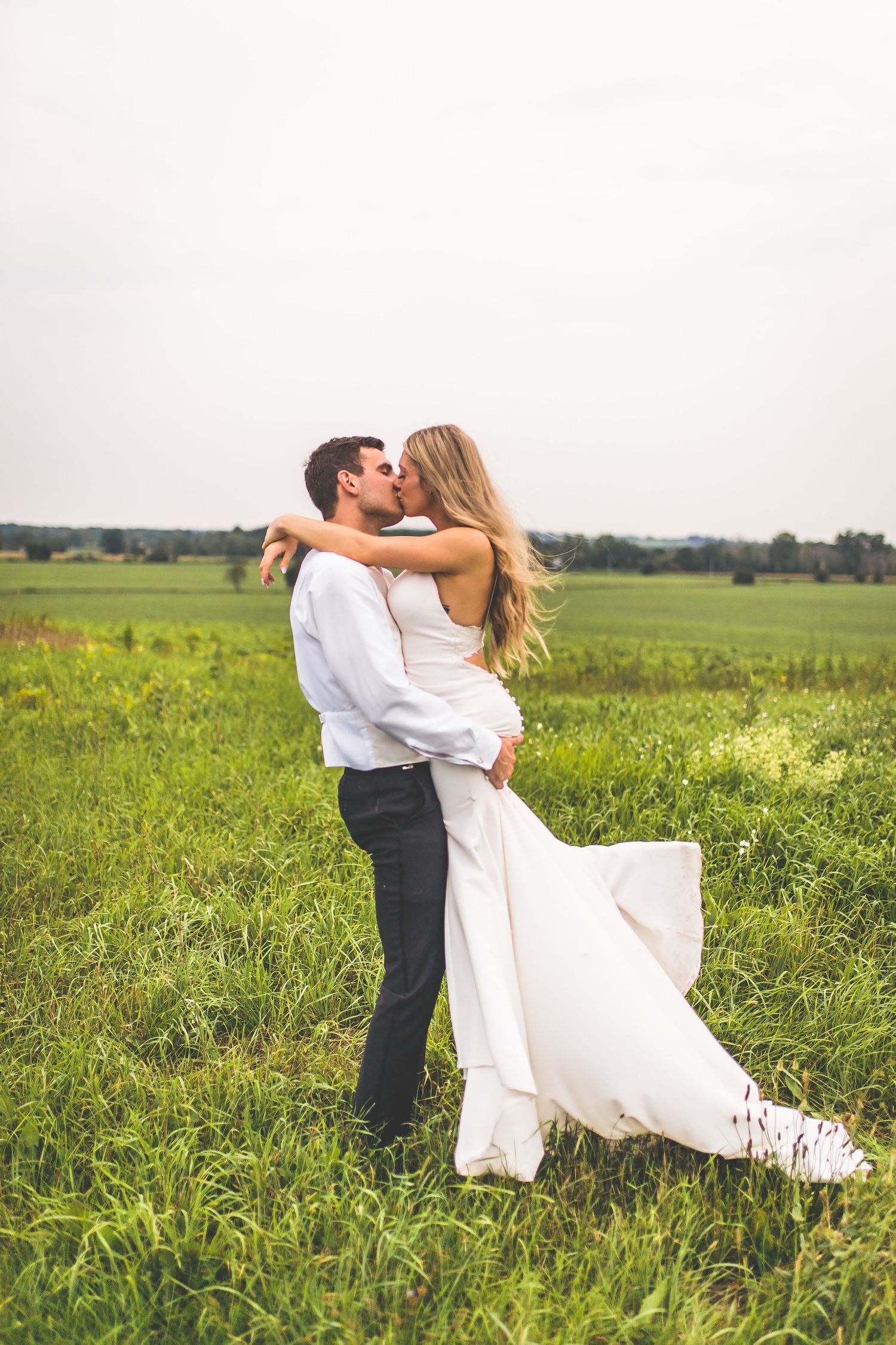 Couple in wedding attire embracing in a scenic meadow with rolling hills in the background.