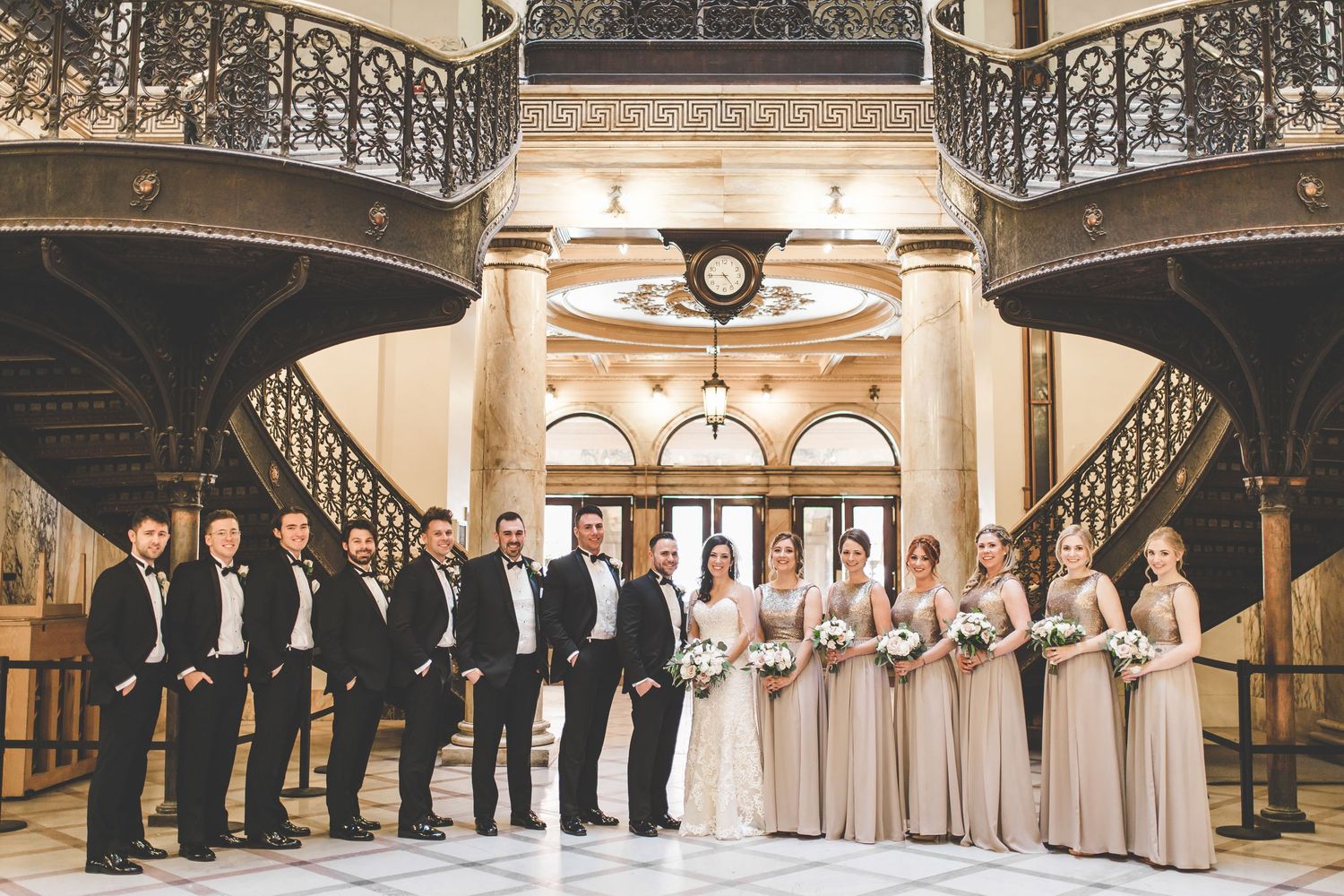Wedding party lined up in an elegant indoor venue with ornate staircase and chandelier lighting.