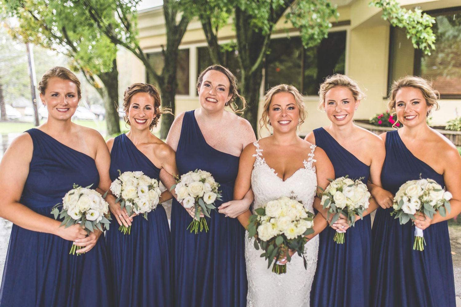 Bridesmaids in navy blue dresses pose with white bouquets outdoors.
