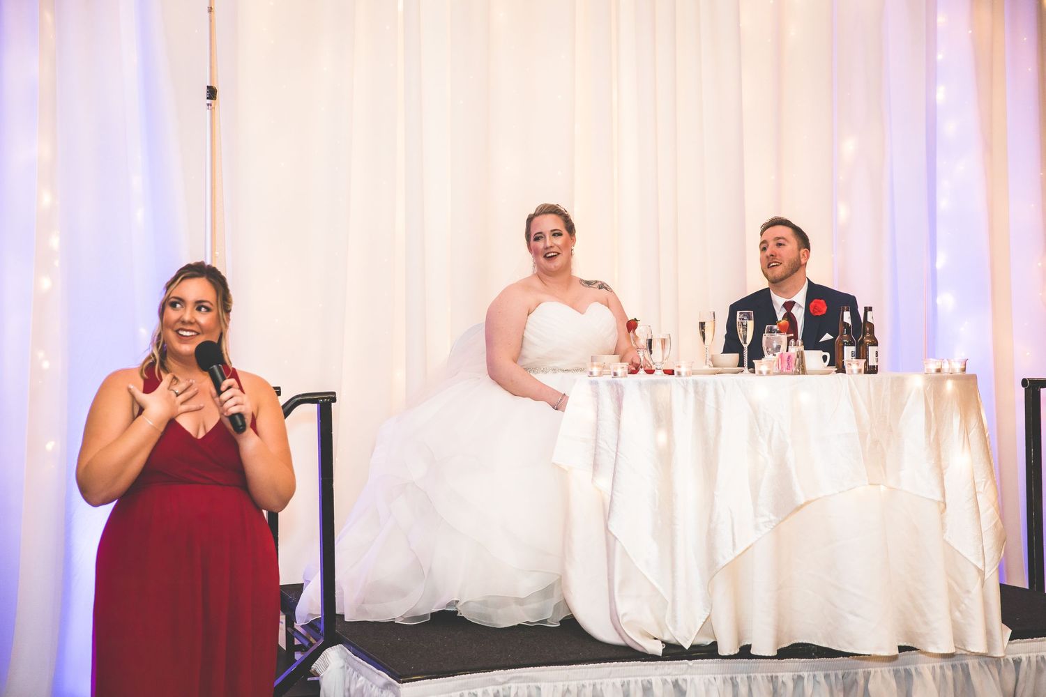 Wedding reception scene with bridesmaid speaking while couple sits at head table.