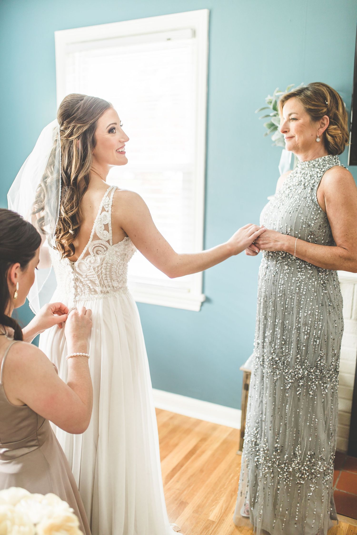 Three women share a joyful moment while getting ready in a blue-walled room with natural light.