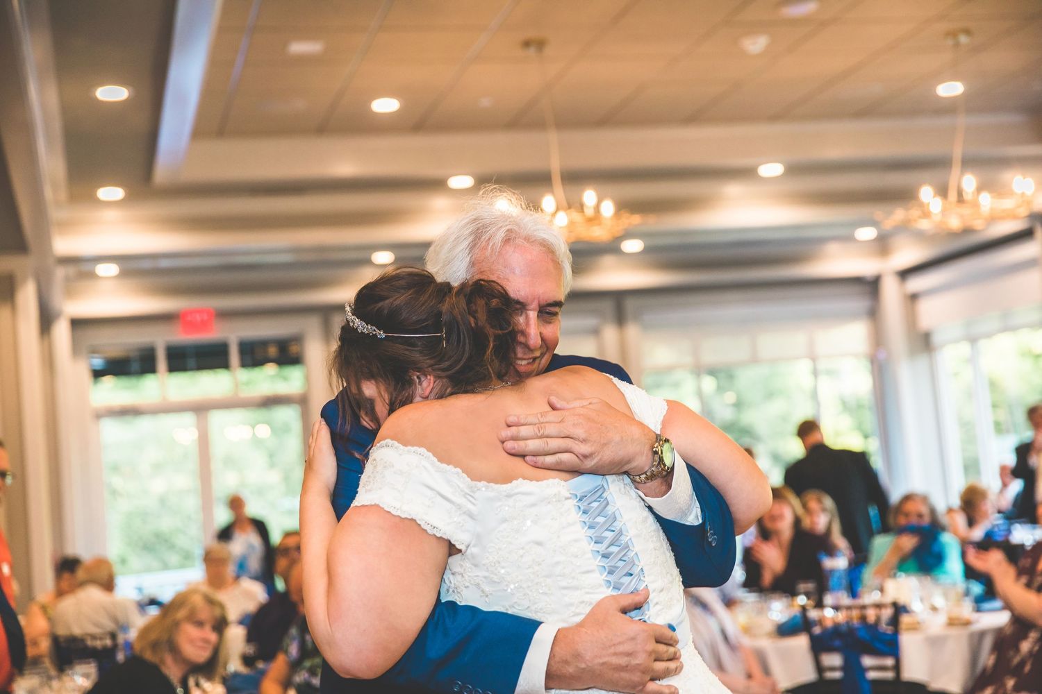 Two people share an emotional embrace during a wedding reception with guests seated in the background.