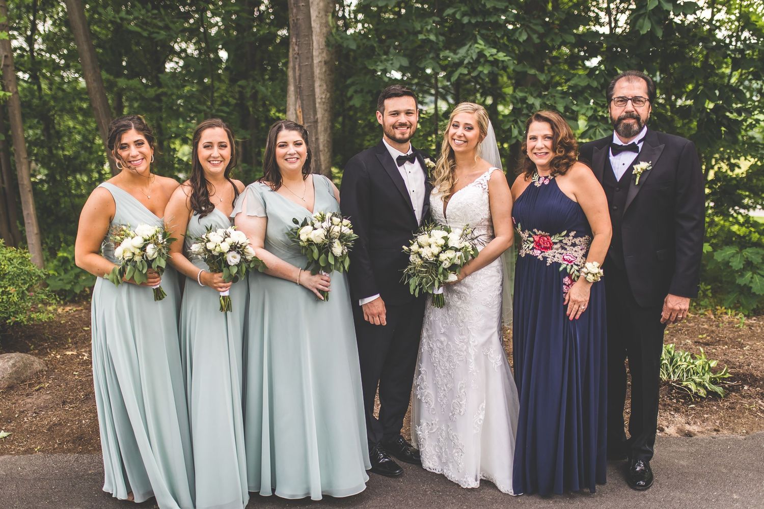 Wedding party poses together in mint green and navy attire against wooded backdrop.