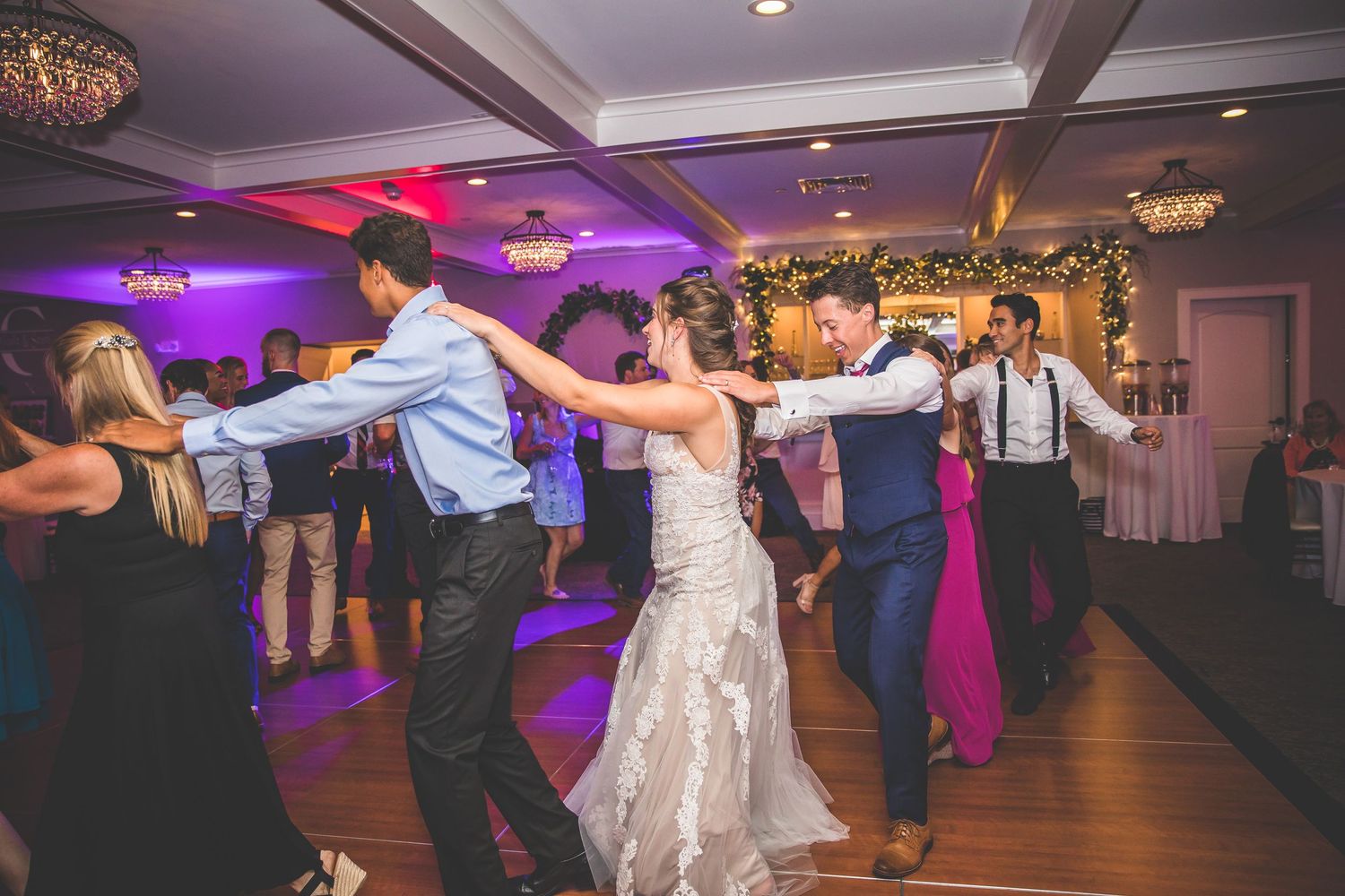 Wedding reception guests dancing together on a dimly lit dance floor with purple and red lighting.