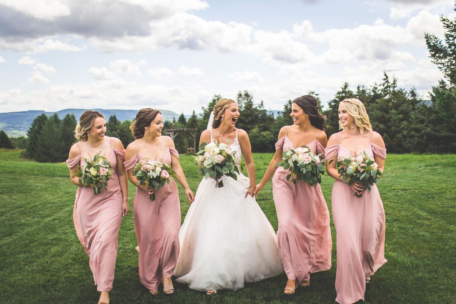 Bridesmaids in blush pink dresses pose with bride in white gown on grassy hillside.