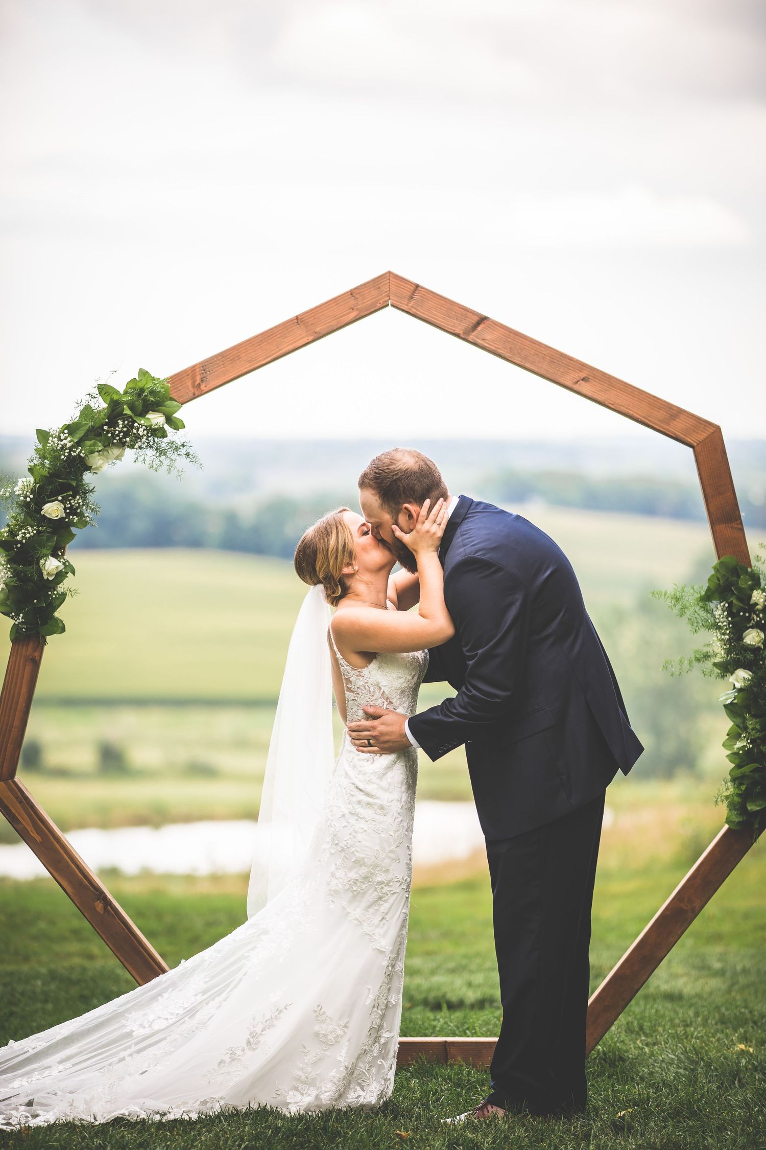 Wedding couple shares romantic kiss under wooden hexagonal arch decorated with greenery against scenic countryside view.
