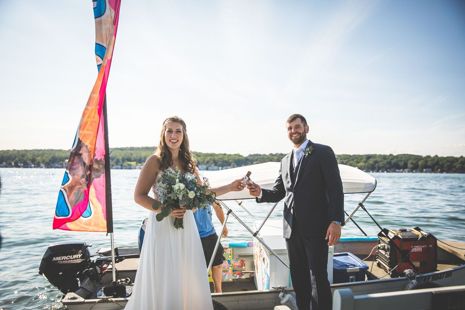 Wedding couple stands on sailboat with colorful sail and lake backdrop.