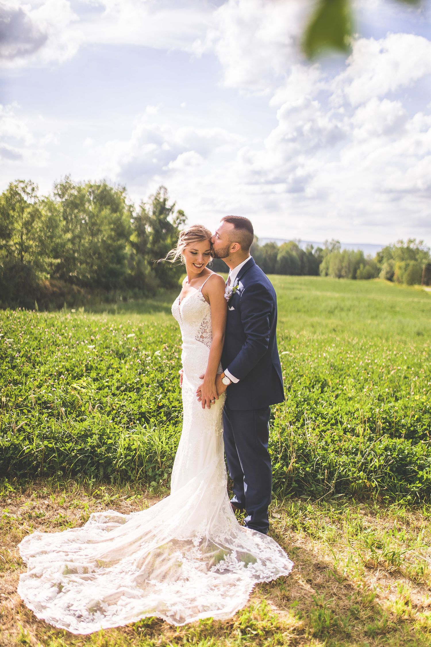 A bride in a lace train dress poses with her groom in a sun-drenched field.