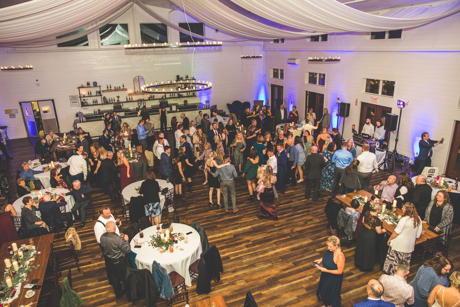 Large wedding reception venue with tables and guests under elegant white draping and blue lighting.
