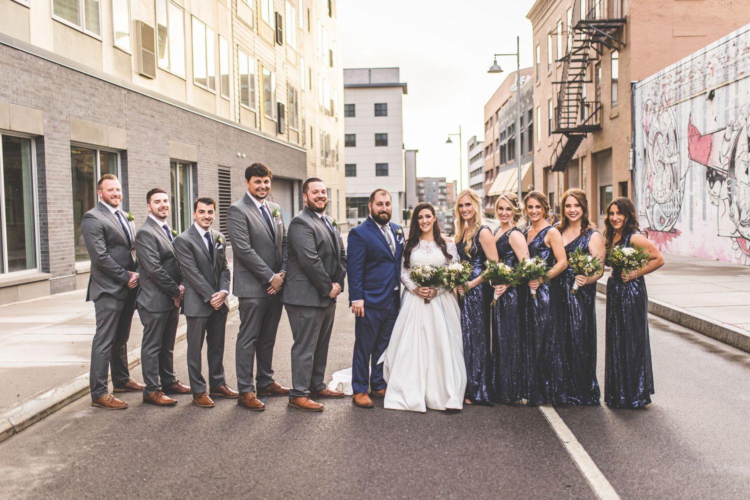 Wedding party poses together on urban street with groomsmen in gray suits and bridesmaids in navy dresses.
