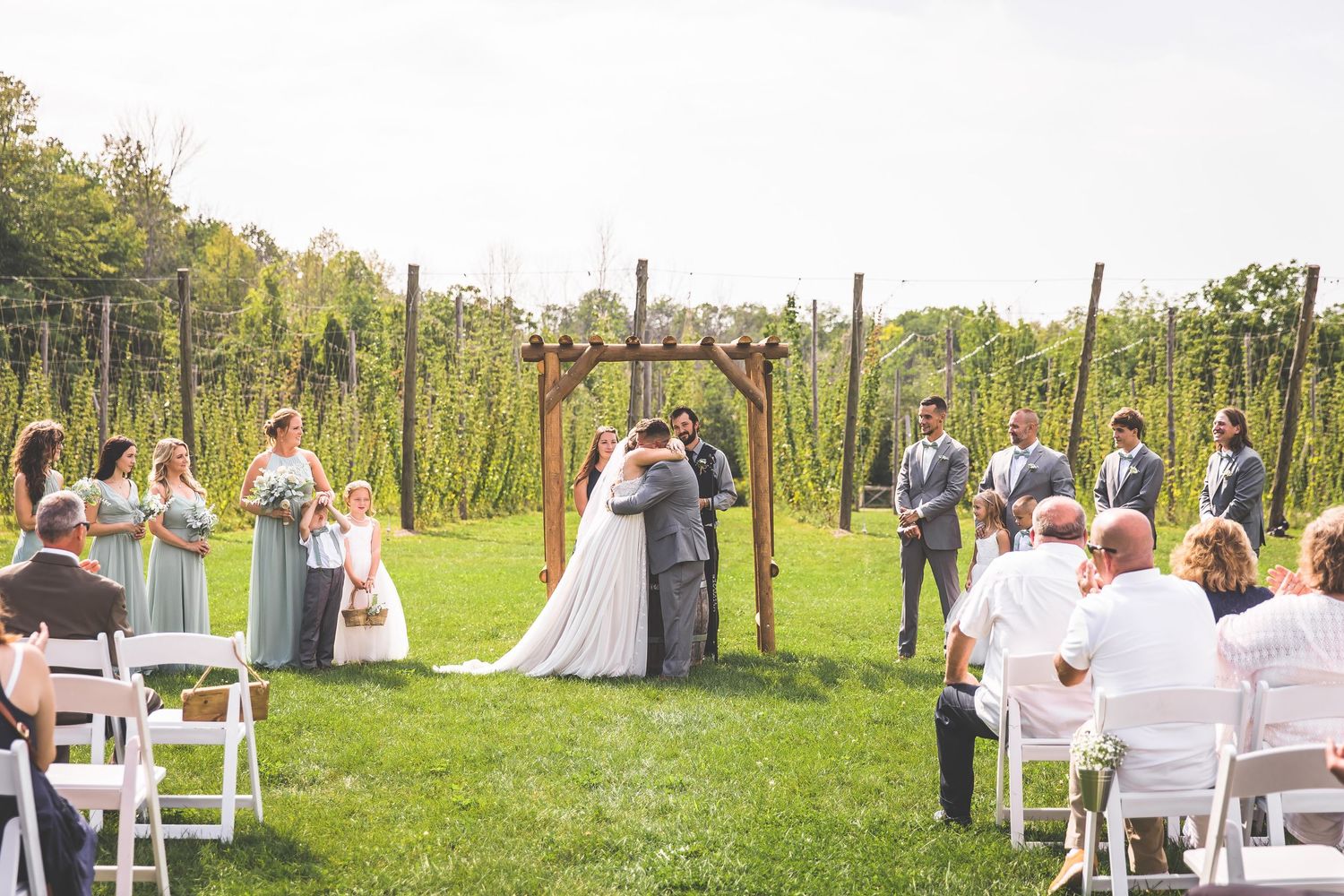 Wide view of outdoor garden wedding with white chairs and wooden pergola setup.