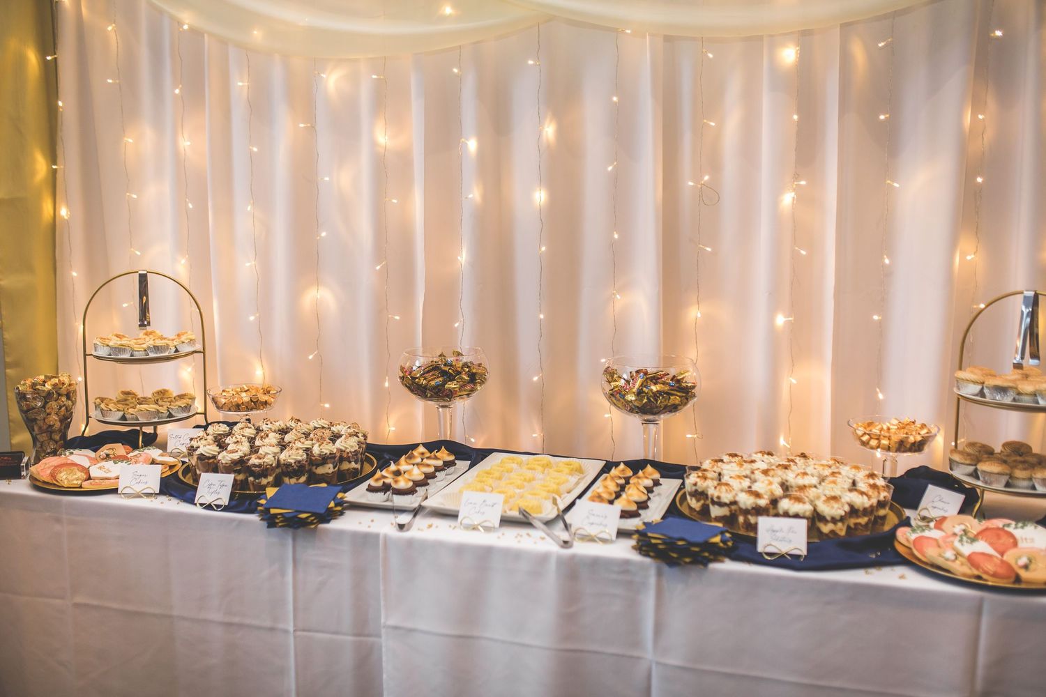 Elegant dessert table display with tiered stands and twinkling fairy light backdrop.