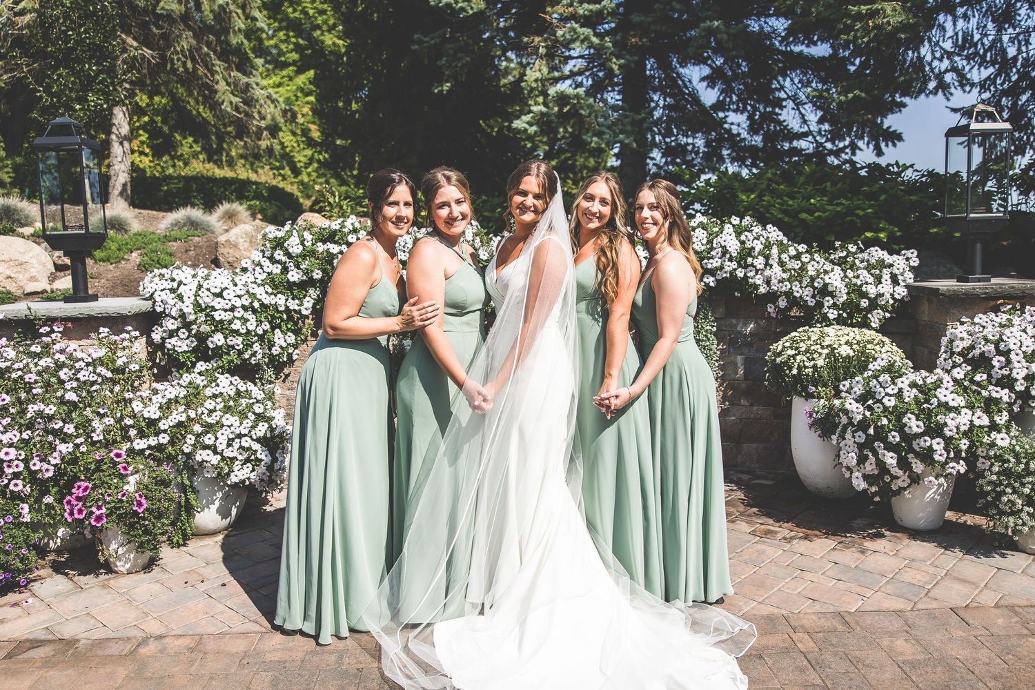 Wedding party in sage green dresses poses against stone wall with floral decorations.