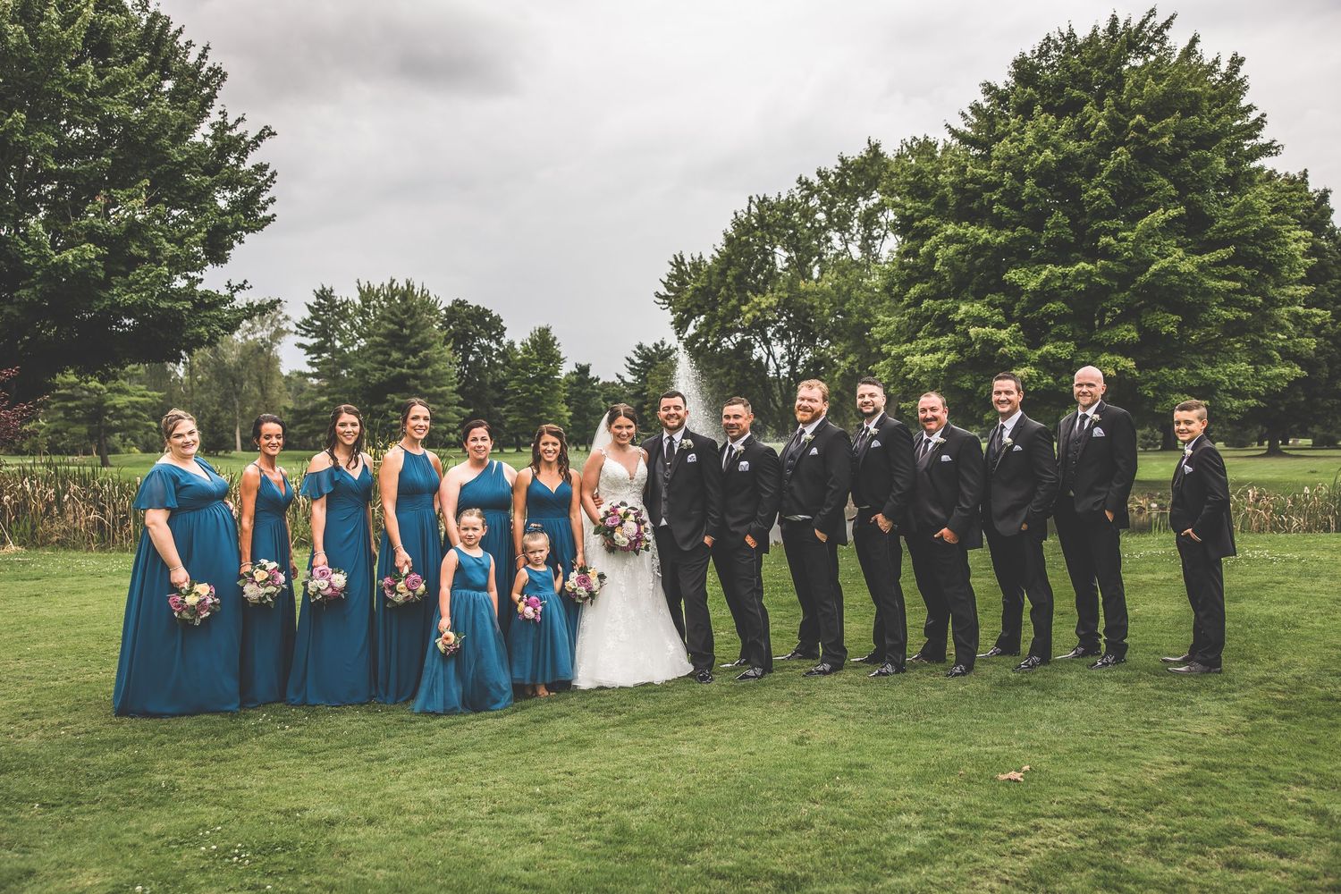 Wedding party in blue dresses and black suits poses on grassy hill for formal photo.