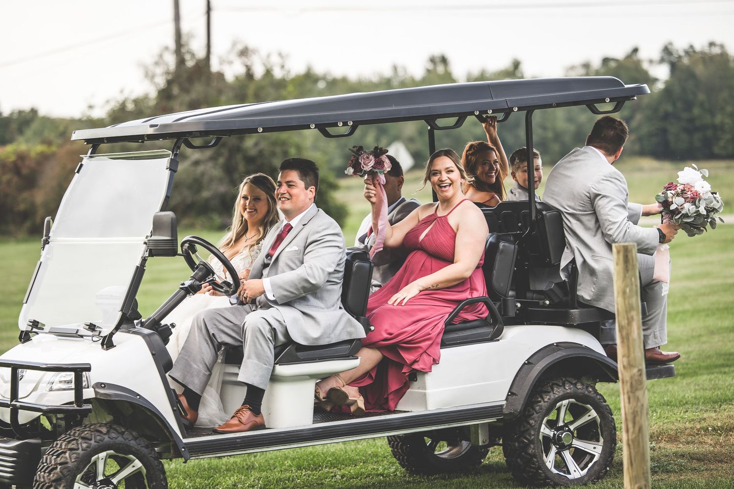 Wedding party rides in vintage golf carts across a lush green course on a sunny day.