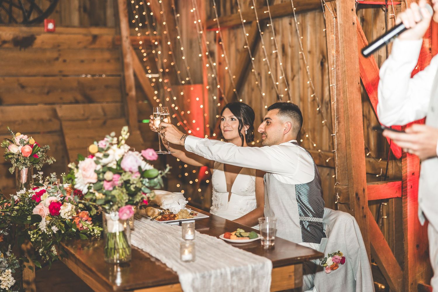 Wedding celebration in a wooden barn venue illuminated by warm string lights and decorated with pink flowers.