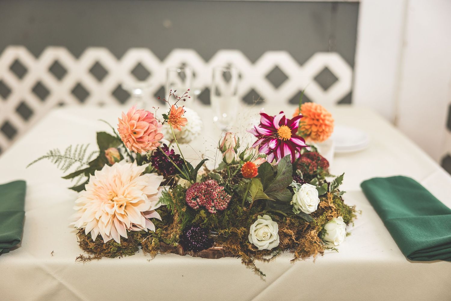 Rustic floral centerpiece with peach dahlias and purple flowers arranged on a white tablecloth with green napkins.