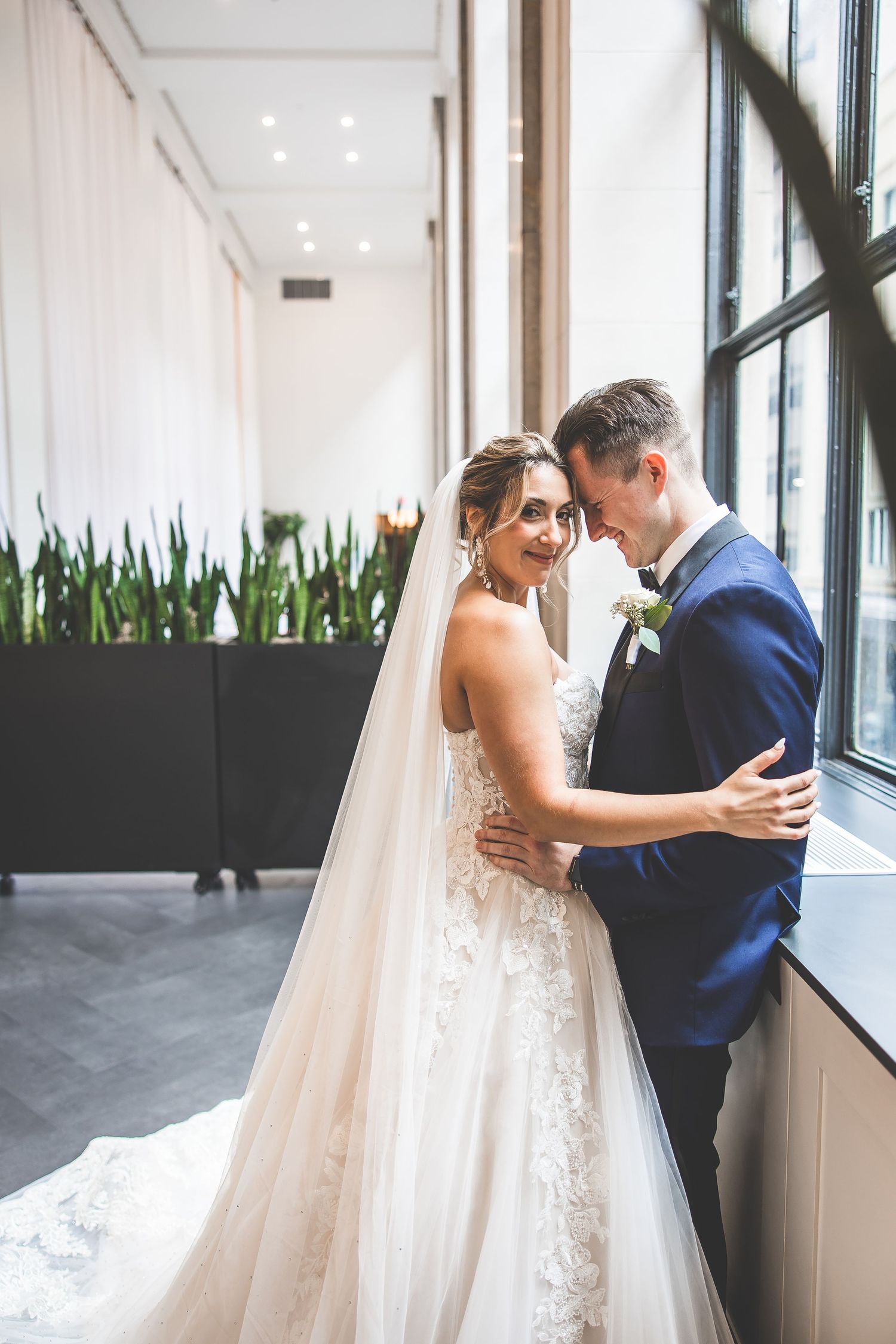 A romantic wedding portrait by large windows with natural light streaming in.