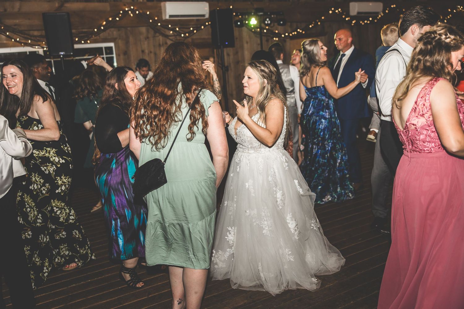 Wedding party dancing in a rustic venue decorated with string lights and wooden beams.