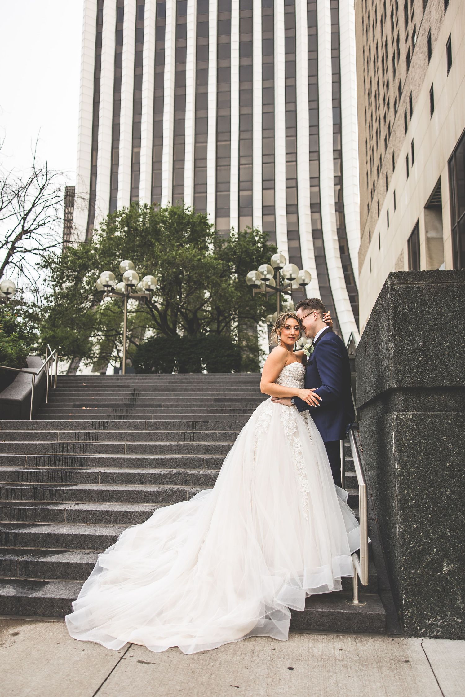 Bride in flowing white gown and groom in navy suit embrace on stone steps near city buildings.