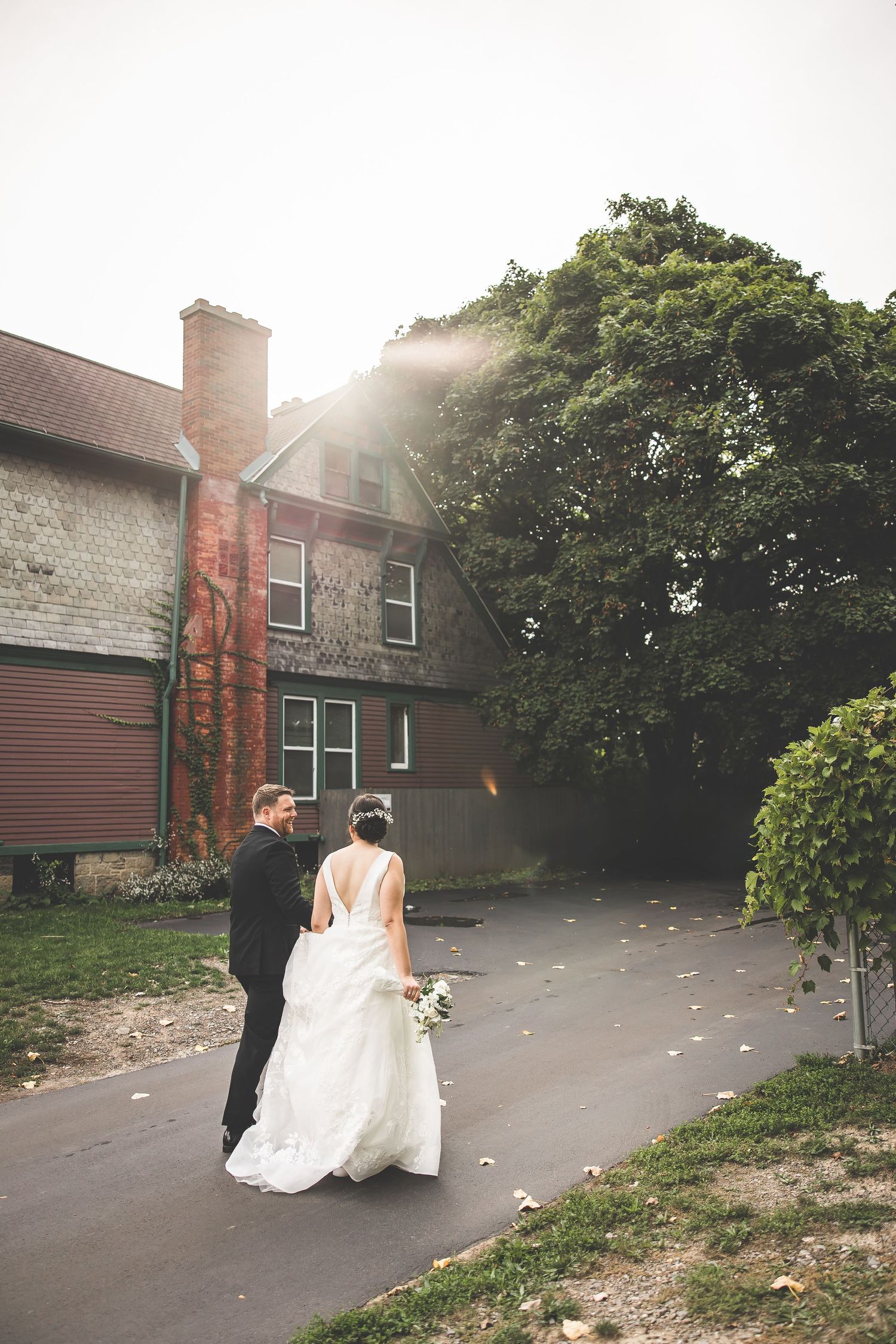 A romantic wedding couple walking together on a garden path with trees and a brick building in the background.