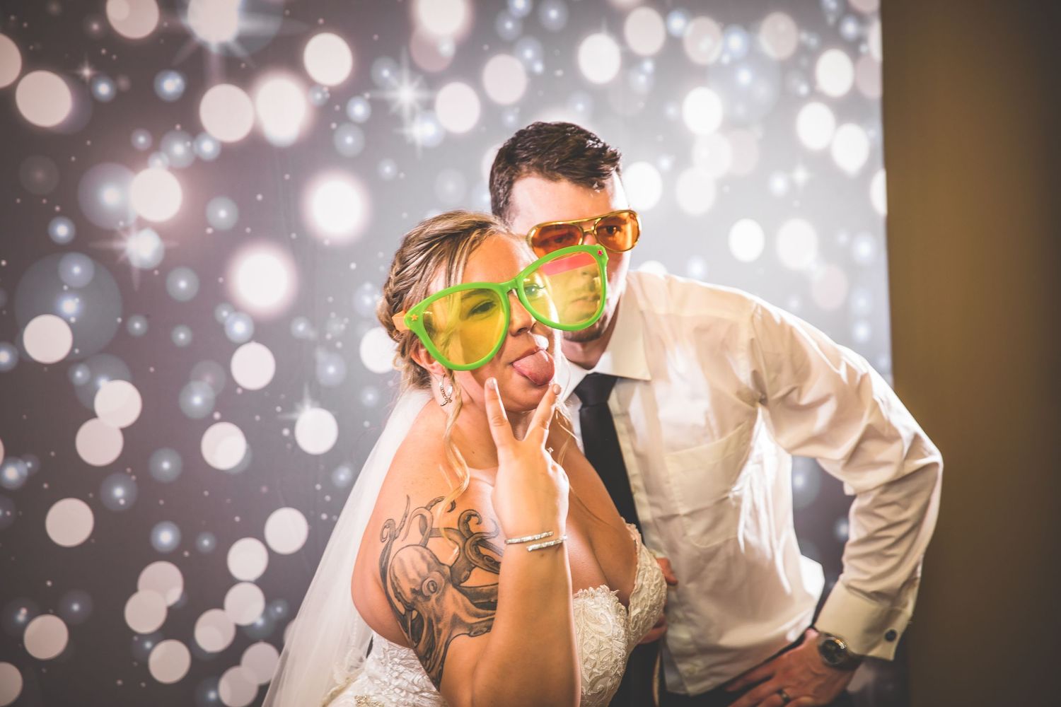 A couple wearing colorful party glasses poses against a sparkly bokeh background.