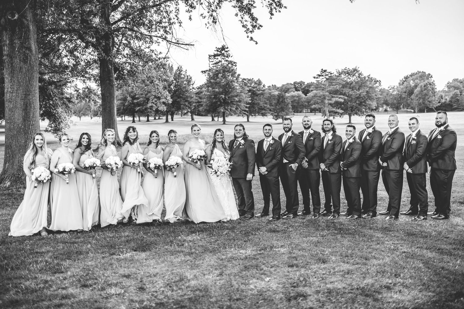 A large wedding party lined up in formal attire under trees in black and white.