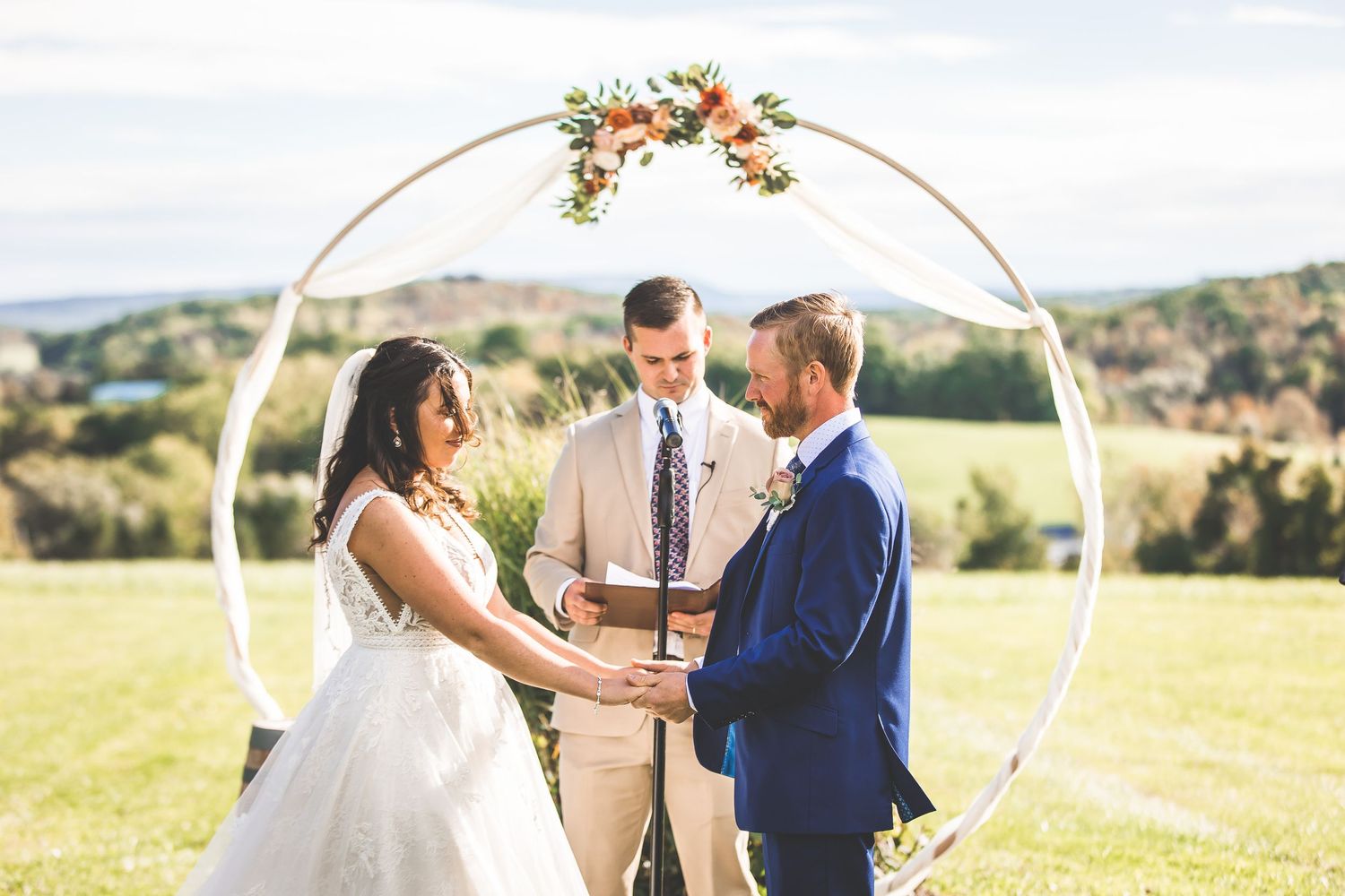 Wedding ceremony under a circular arch decorated with flowers and fabric in an outdoor hilltop setting.