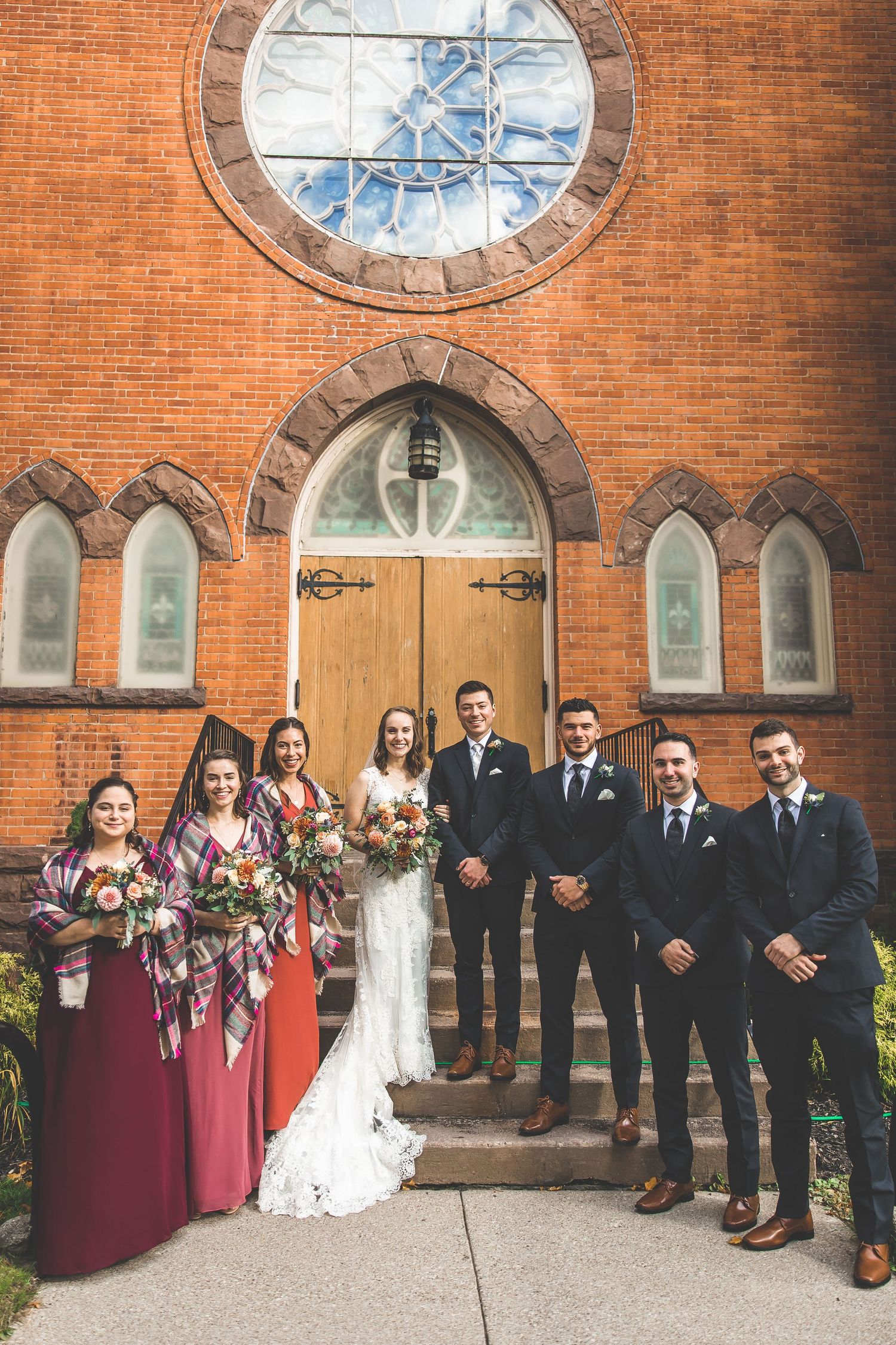Wedding party lined up outside brick church with gothic windows and burgundy bridesmaid dresses.