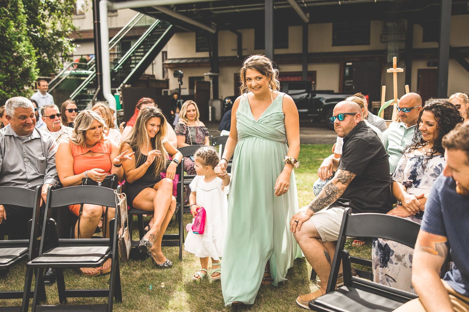 Guests gathered at outdoor wedding venue with covered structure and greenery.