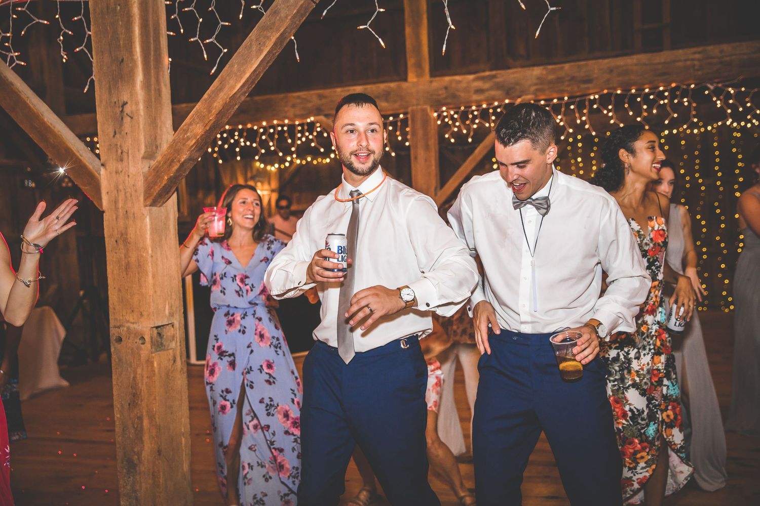 Wedding guests dancing and celebrating at a rustic barn reception with string lights and wooden beams overhead.