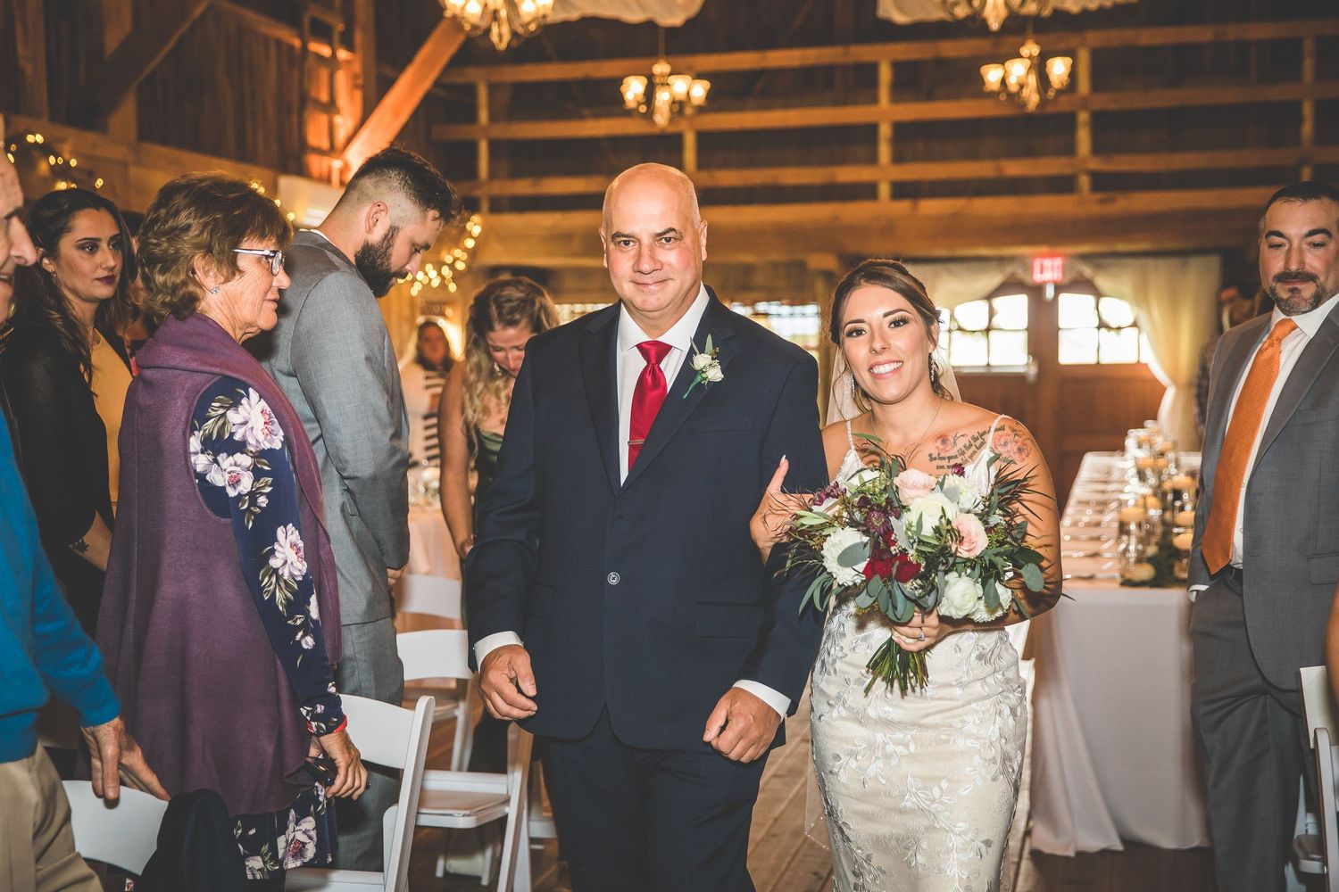 Wide angle view of wedding ceremony in rustic barn with chandelier lighting and floral decorations.