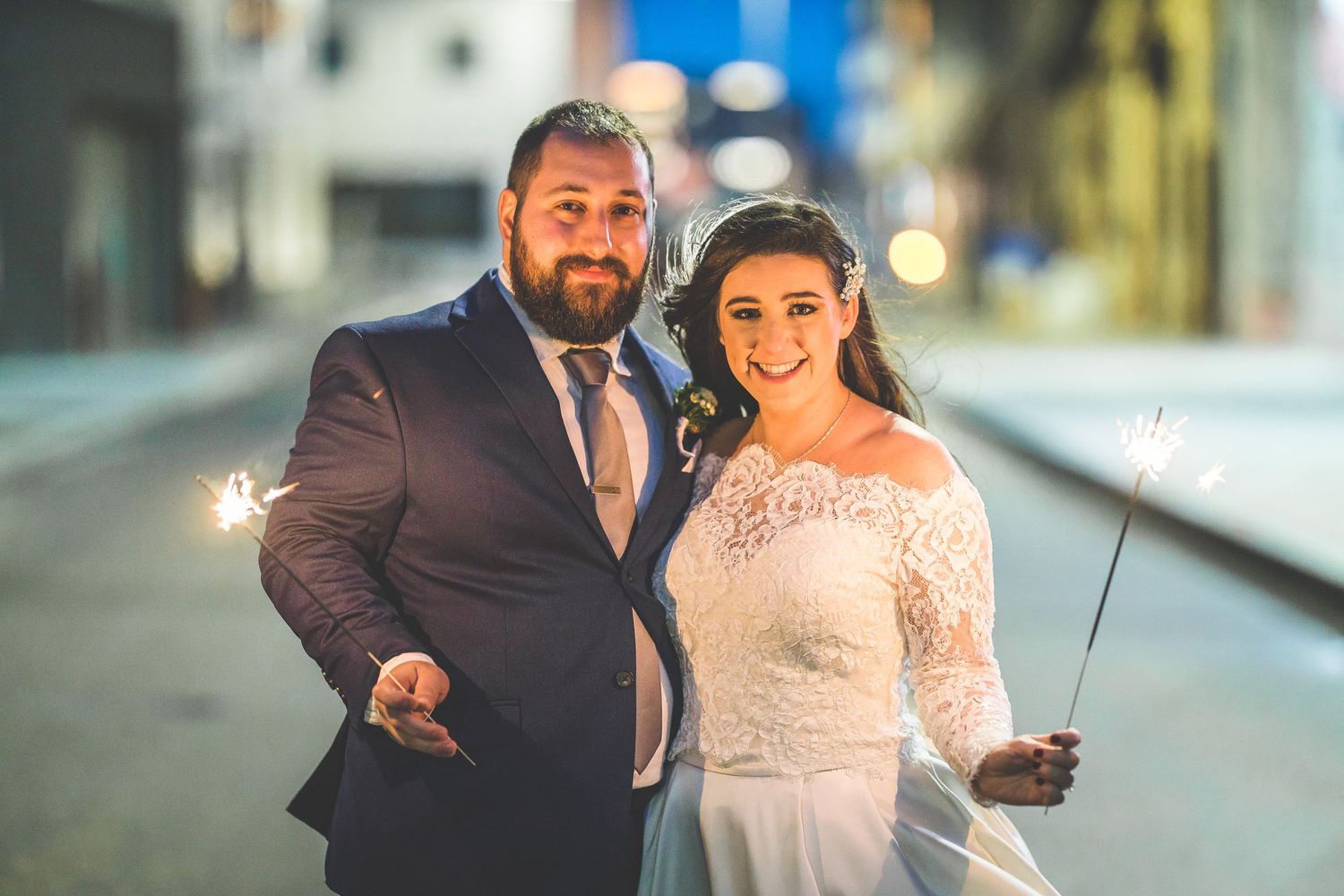 Couple holds sparklers while posing on city street at night during wedding celebration.