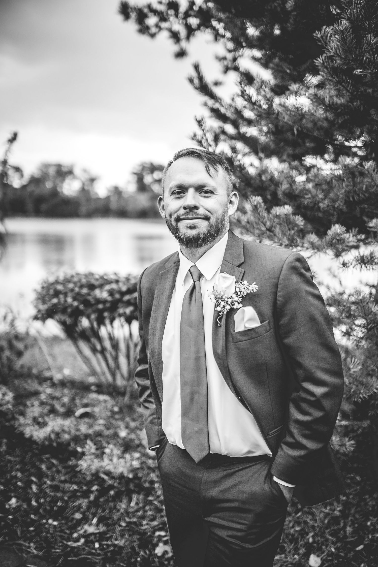 Black and white portrait of groom in suit standing by lake with natural landscape background.
