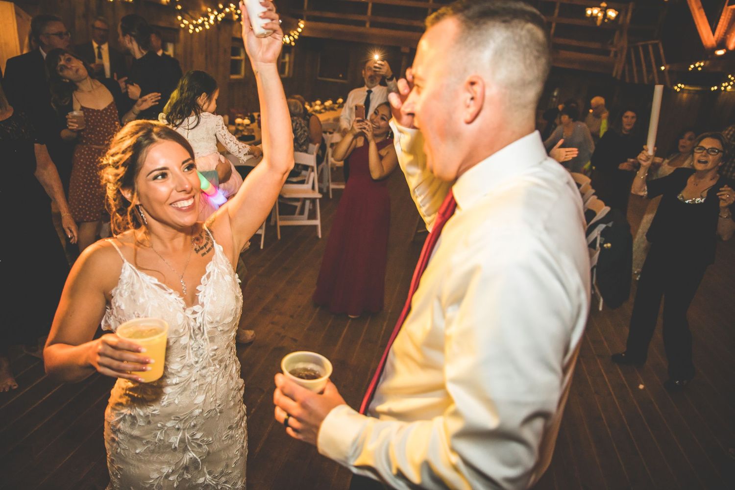 Wedding guests dancing and celebrating on a rustic wooden dance floor with warm lighting and chandeliers.