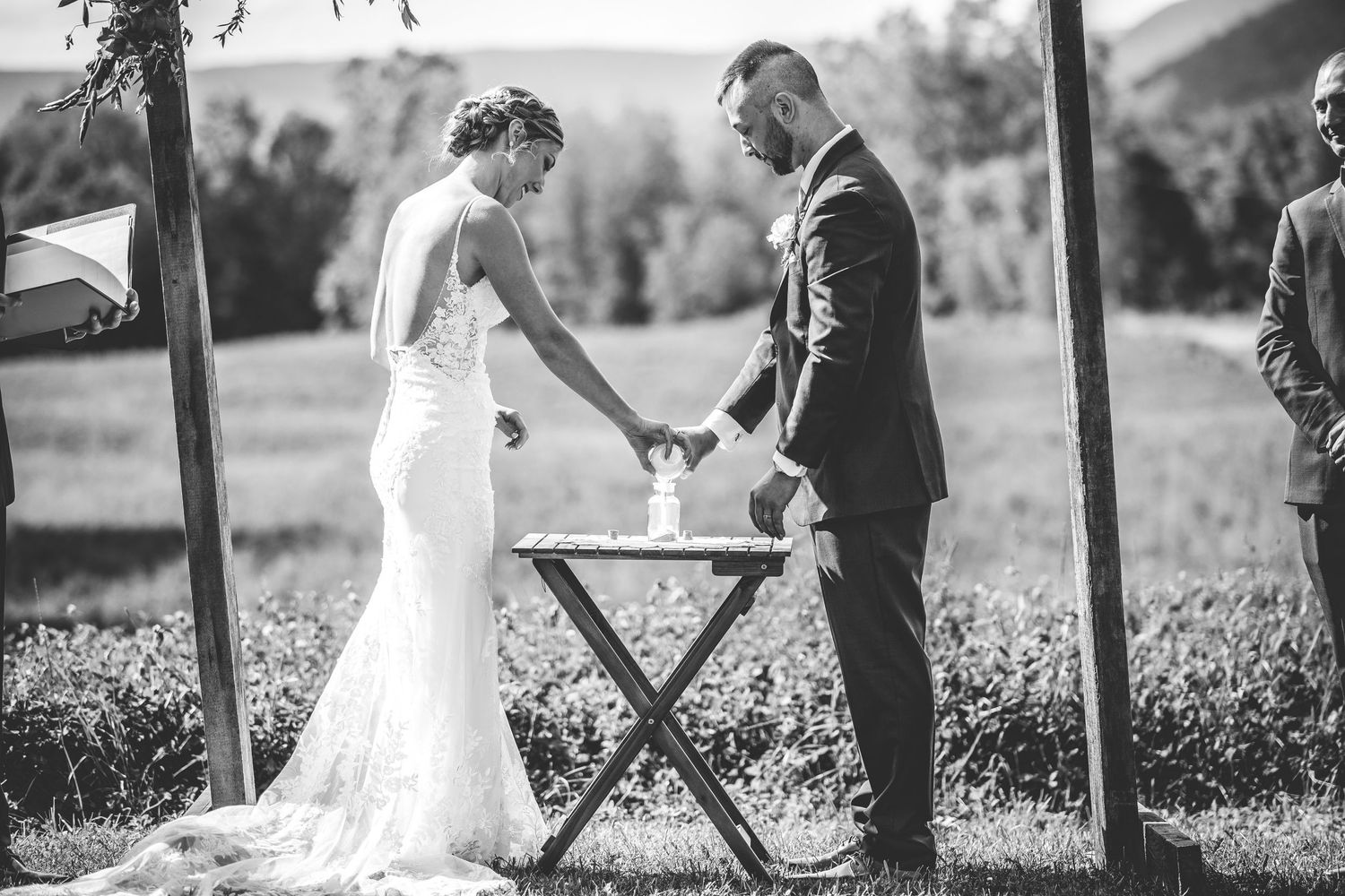 Black and white photo of wedding ceremony under wooden arch in natural outdoor setting.