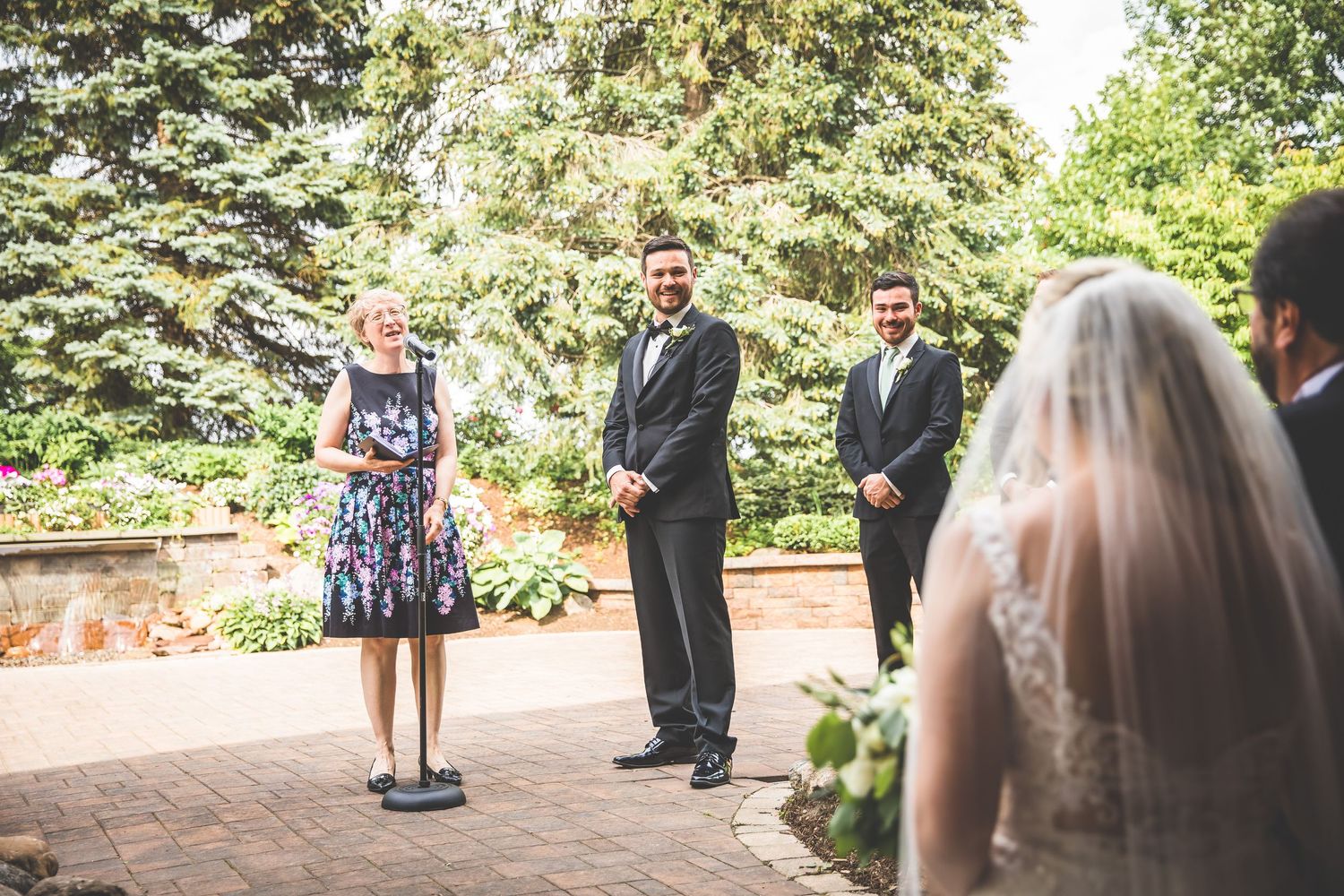 Outdoor wedding ceremony with attendants standing on a brick patio surrounded by greenery.